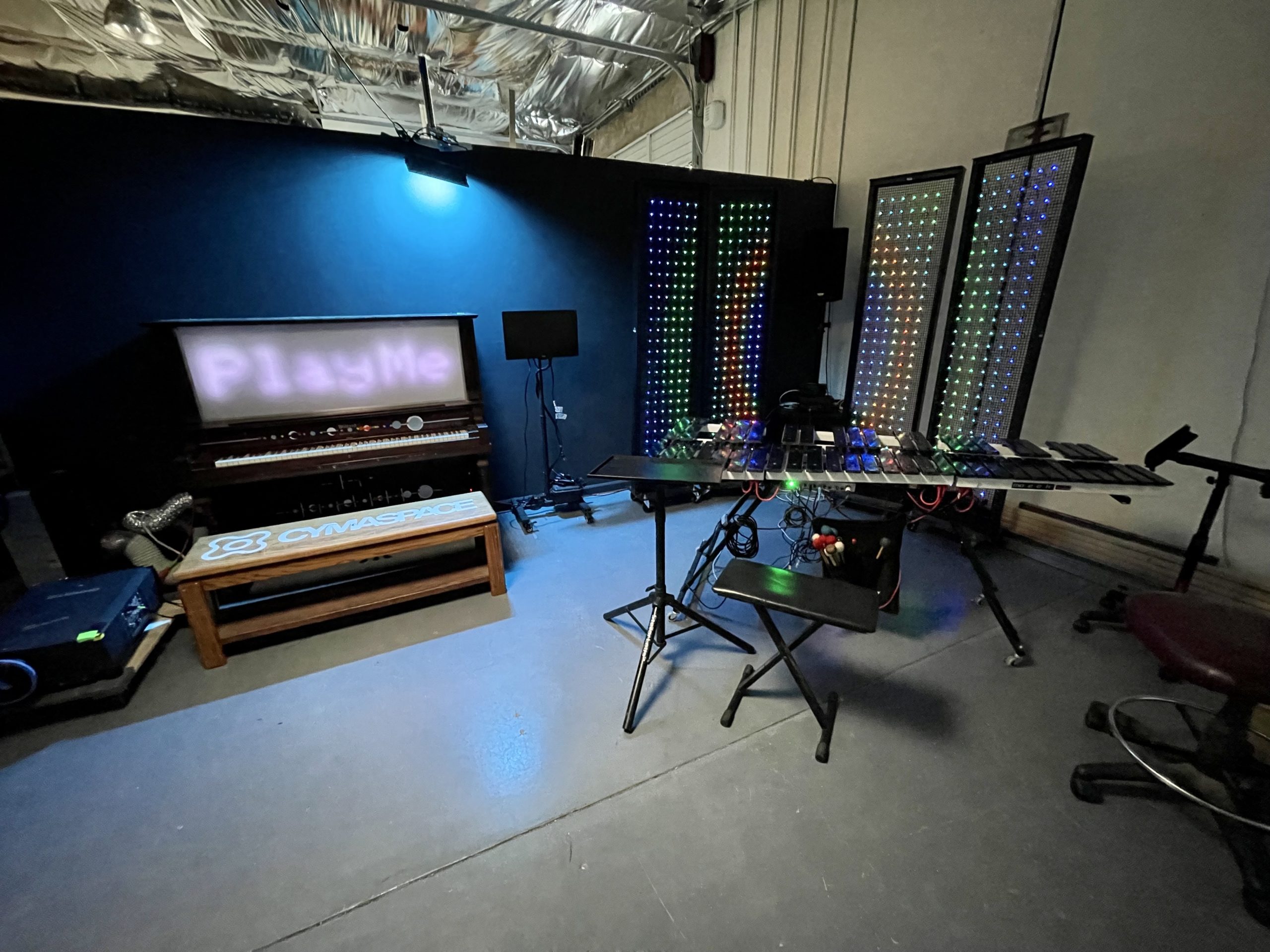 a corner of a workshop with a dark coal gray wall backdrop, there is an upright piano with a display that says "Play Me", next to it are four light fixtures with LED pixels displaying rainbow colors in a circular pattern of blue, green, orange, blue. In front of the light fixtures is an electronic marimba instrument with black keys.